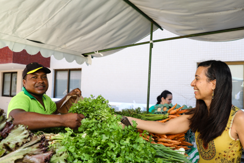 Fotografia contendo uma barraca de feira, com cenoura e tipos de alfaces, um vendedor e uma cliente 