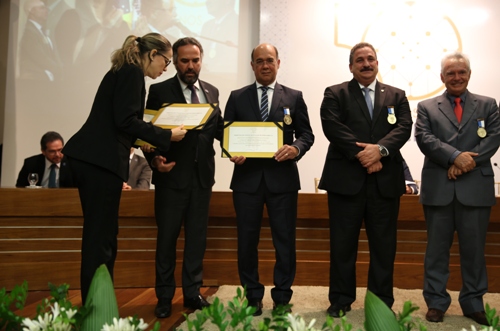 Fotografia de autoridades com roupas formais, em cima de um palco, recebendo diplomas de homenagem