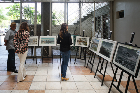 Foto do hall do edifício-sede do TRT6 com os quadros expostos e três pessoas olhando as imagens que estão em quadros pendurados em cavaletes
