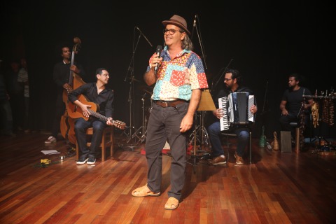 Fotografia de Hélio Donato cantando (veste calça cinza, camisa de retalhos coloridos e chapéu), ao fundo banda com Teles Jr ao violão e outros músicos no contra-baixo, bateria, acordeom e percussão