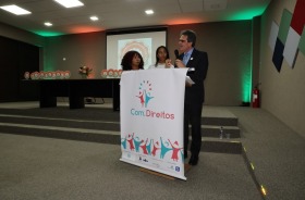 Desembargador Ruy Salathiel fala em púlpito próximo a duas jovens aprendizes que olham para ele