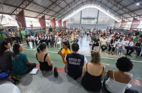 Na foto uma mulher fala para dezenas de pessoas sentadas numa quadra de esportes