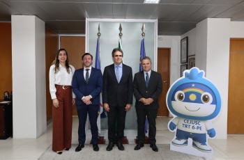 Quatro pessoas posam em frente a bandeiras ao lado do mascote do CEJUSC TRT-6.