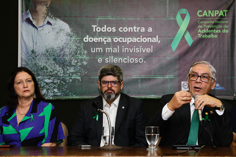 Fotografia de três autoridades sentadas à mesa, uma delas fala ao microfone. Ao fundo um banner com os dizeres “Todos contra a doença ocupacional, um mal invisível e silencioso. Canpat – Campanha Nacional de Prevenção de Acidentes do Trabalho”