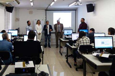 Fotografia de uma sala de aula. Os quatro professores aparecem de frente junto a uma tela de projeção. Os alunos estão sentados, cada um em uma mesa com computador 