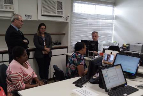 Fotografia de pessoas dentro de uma sala de audiência. Uma delas conversa - via videoconferência – com outra pessoa que aparece na tela do computador