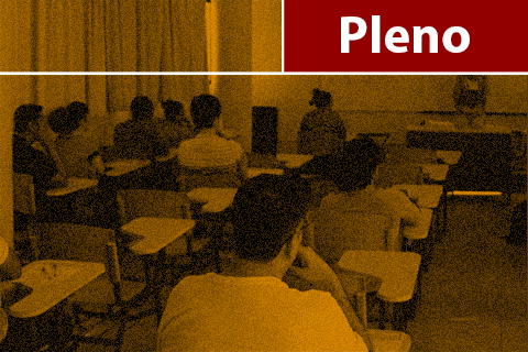 Fotografia, com filtro amarelo, de uma sala de aula – alunos sentados nas carteiras e pessoa à frente da sala, próximo à mesa do professor – texto “Pleno” na lateral superior esquerda.