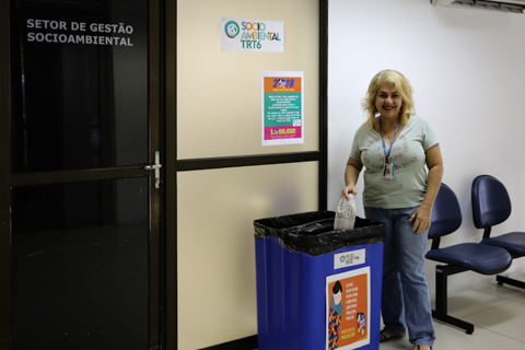 Fotografia de uma mulher depositando uma meia em um coletor. É possível ler cartaz com o texto &quot;Socioambiental&quot; e letreiro de porta &quot;Setor de Gestão Socioambiental&quot;&quot;