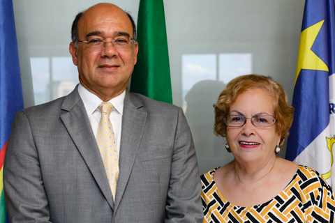 Fotografia, tipo retrato, de duas pessoas. Ao fundo, as bandeiras do Brasil, de Pernambuco e do Recife