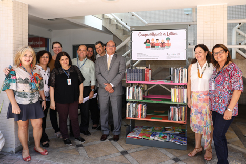 Grupo de pessoas, em pé, posando para foto, ao lado de uma estante de livros, onde há um banner escrito “Compartilhando a leitura”. Todos estão em ambiente externo.