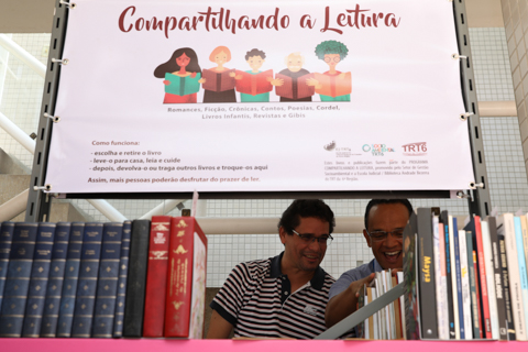 Dois servidores observam livros em estante. Acima deles, há um banner escrito “Compartilhando a leitura”