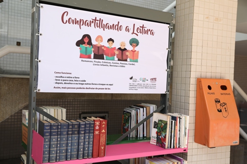 Fotografia de uma estante com livros. No topo há um banner com ilustração representando pessoas lendo e com o texto &quot;Compartilhando a Leitura&quot;