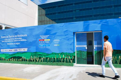 Fotografia de um muro pintado em azul, com símbolo da coleta seletiva e texto “Posto de Troca de Resíduos”. Há outros textos e imagens que não são possíveis ver em detalhe. Uma pessoa caminha próxima ao muro