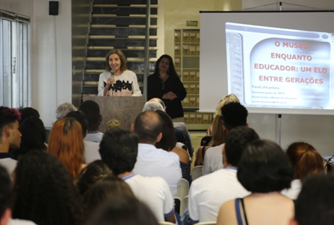  Fotografia da desembargadora Eneida Melo discursando em um púlpito. No plano de funda, a servidora Marcília Gama