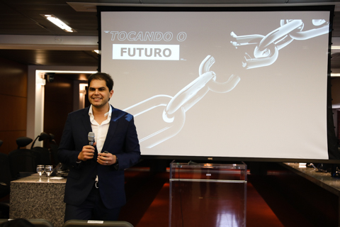 Fotografia do palestrante, em pé, posicionado a frente de um telão, onde se lê a projeção &quot;Tocando o futuro&quot;