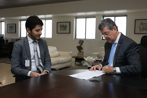 fotografia de dois homens em trajes formais, sentados a mesa de reuniões. Um assina um documento enquanto o outro observa