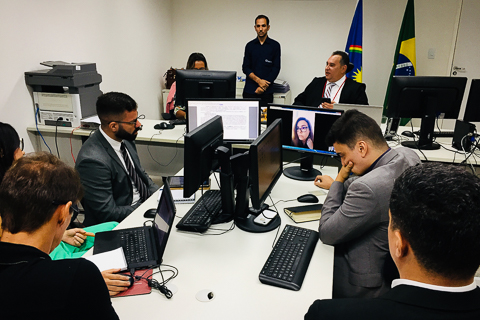 Fotografia de pessoas participando de audiência. ?Na tela do computador é possível ver uma mulher participando por videoconferência