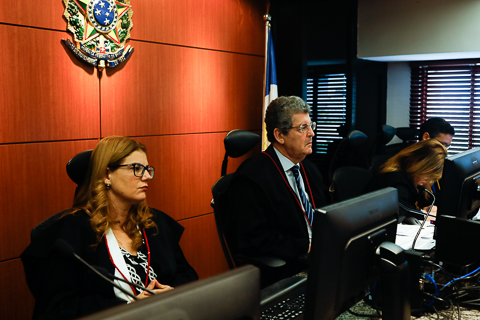 Fotografia de três pessoas de toga, sentadas à frente de computadores. Atrás dela é possível ver o Brasão da República