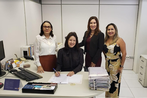 Fotografia de quatro mulheres em trajes profissionais em uma sala de trabalhos. Uma delas assina um documento
