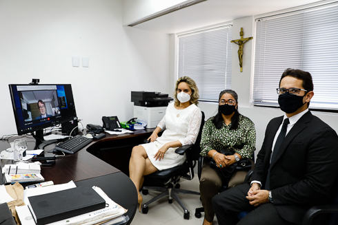 Participantes da reunião sentados em frente ao computador em que aparece o participante via teleconferência