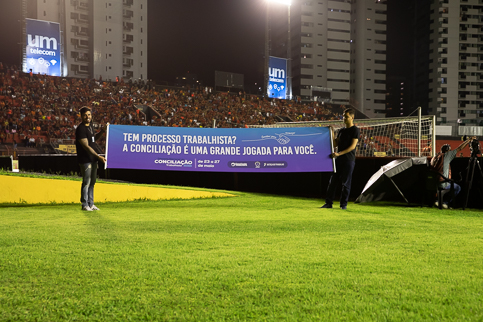 Foto de duas pessoas segurando uma faixa de divulgação da Semana de Conciliação em frente à torcida