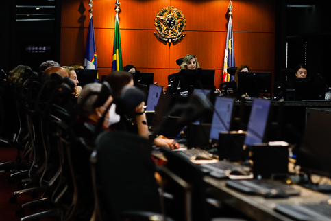 Foto dos desembargadores sentados em frente a computadores. A presidente está na parte central da sala, próxima as bandeiras do Brasil e de Pernambuco e ao Brasão da República