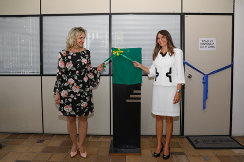 Fotos das magistradas Nise Pedroso e Tânia Chenk Allatta puxando fita para remover tecido que cobre a placa de inauguração da vara