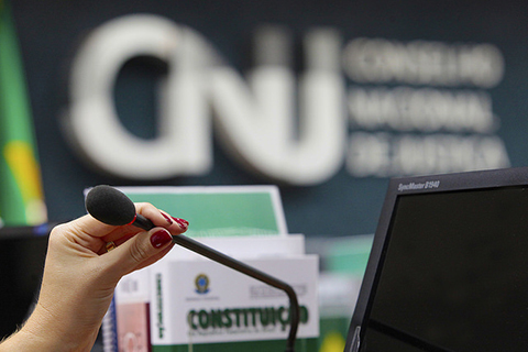 Fotografia da mão de uma mulher junto a um computador e a um microfone, ambos utilizados em audiência judicial. Ao fundo a logomarca do CNJ.