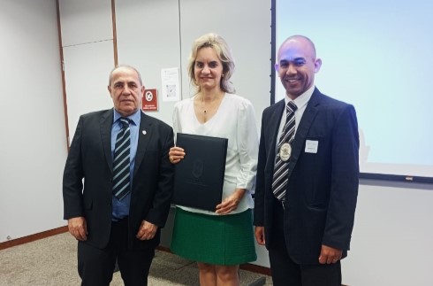 presidente Nise Pedroso ladeada por dois homens de ternos. Todos estão dentro de um auditório