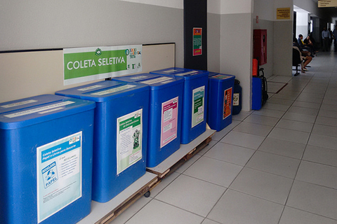 Sequência de coletores em cor azul. Acima deles, cartaz escrito “Coleta Seletiva”. Ao fundo, grande corredor, onde se vê pessoas andando e outras sentadas em logarina.