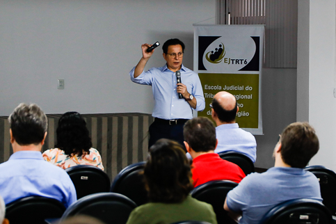 foto de palestrante ao fundo falando ao microfone e segurando passador de slides; plateia de costas no primeiro plano