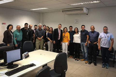 Fotografia de 16 pessoas, em pé, olhando para a câmera, dentro de uma sala de trabalho