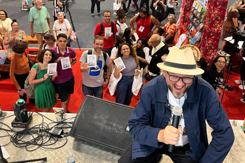 Foto do servidor autor do cordel em cima de um palco com pessoas em baixo do palco com o cordela na mão