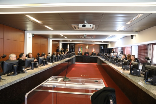 Auditório com mesa em formato de U. Ao longo dela, pessoas estão sentadas em frente a telas de computador