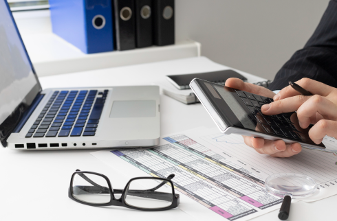 Foto de uma mesa de trabalho com computador e óculos. Uma mão masculina está mexendo em uma calculadora.