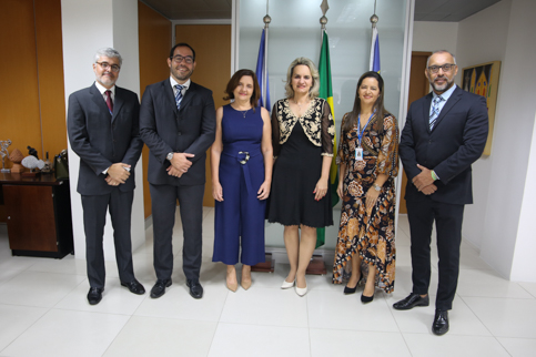 fotografia de seis pessoas em uma sala de reuniões ampla e iluminada. Elas posam à frente das bandeiras do Brasil, Pernambuco e Recife