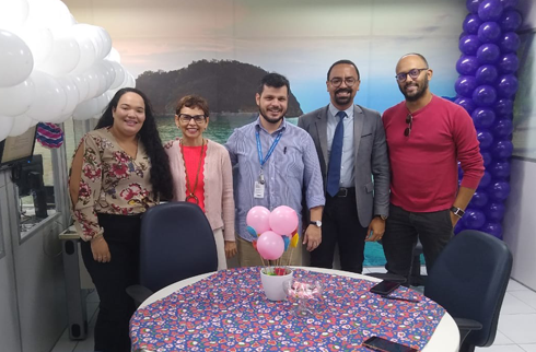 fotografia de cinco pessoas em pé, dentro de uma sala de reuniões. A sala está enfeitada com balões