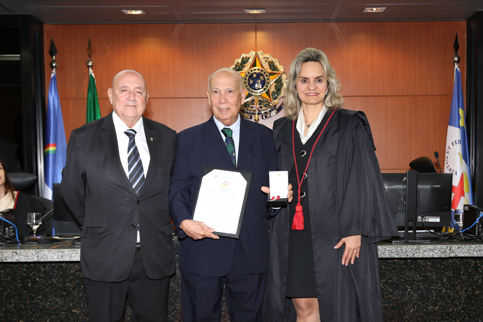 retrato de três pessoas em trajes formais, dentro de um auditório. O homem que está no meio segura uma medalha e um diploma 
