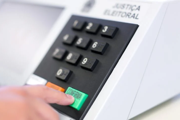 Imagem de uma urna eleitoral com um dedo apertanto a tecla &quot;Confirma&quot;