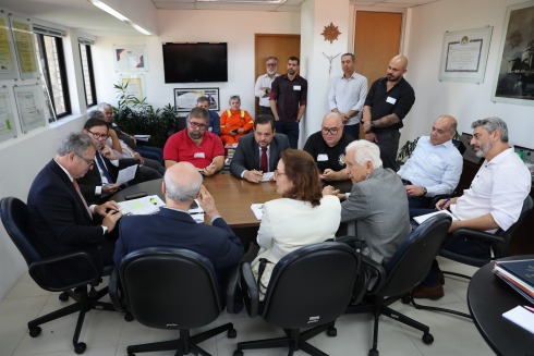 Foto mostra, ao centro, uma mesa de reunião com 10 pessoas sentadas ao redor