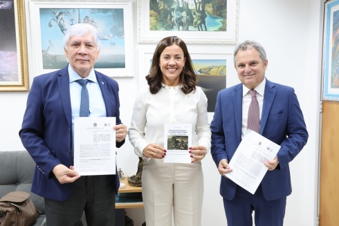 Três pessoas, em escritório com quadros, seguram documentos e um livro. Todos vestem trajes formais.