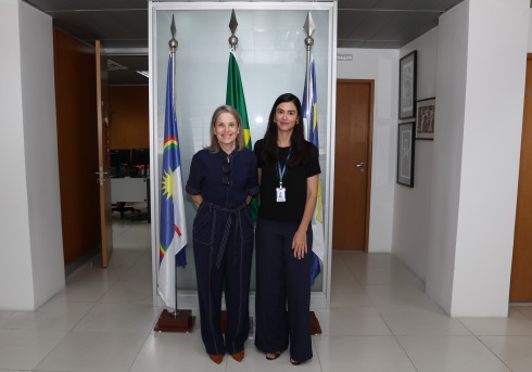 Duas mulheres posam sorrindo em frente a três bandeiras em ambiente institucional