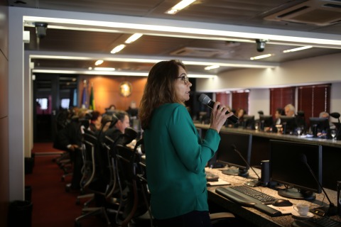 Fotografia de uma mulher falando ao microfone. Ao fundo há uma sala ampla, com autoridades de toga sentadas a frente de computadores