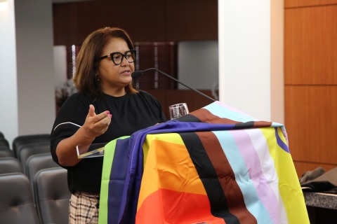 Foto da juíza Ana Cristina falando no púlpito. Há uma bandeira da causa LGBTQIA+ em destaque