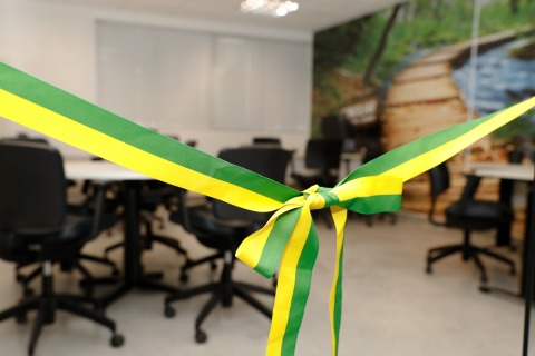 Fotografia com foco em uma fita de inauguração e, por trás, desfocada, uma sala com várias mesas e cadeiras