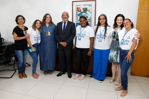 fotografia de pessoas em pé dentro de uma sala de reuniões, algumas usam camisa de campanha da coleta seletiva