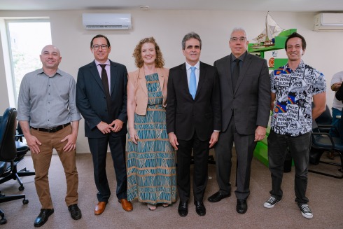 Foto mostra seis pessoas adultas, em pé, lado a lado, em uma sala de parede bege