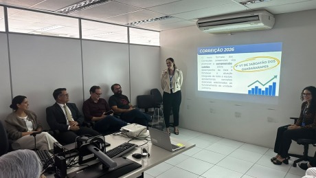 Na foto, aparece uma mulher em pé, fazendo uma aparesentação, e 5 adultos sentados