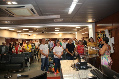 foto de pessoas em pé dentro de um auditório. No palco uma professora fala ao microfone. O local possui alguns enfeites de Carnaval, sobrinha e estandarte