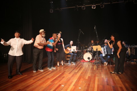 Fotografia de cinco pessoas cantando em cima de um palco, ao fundo, banda